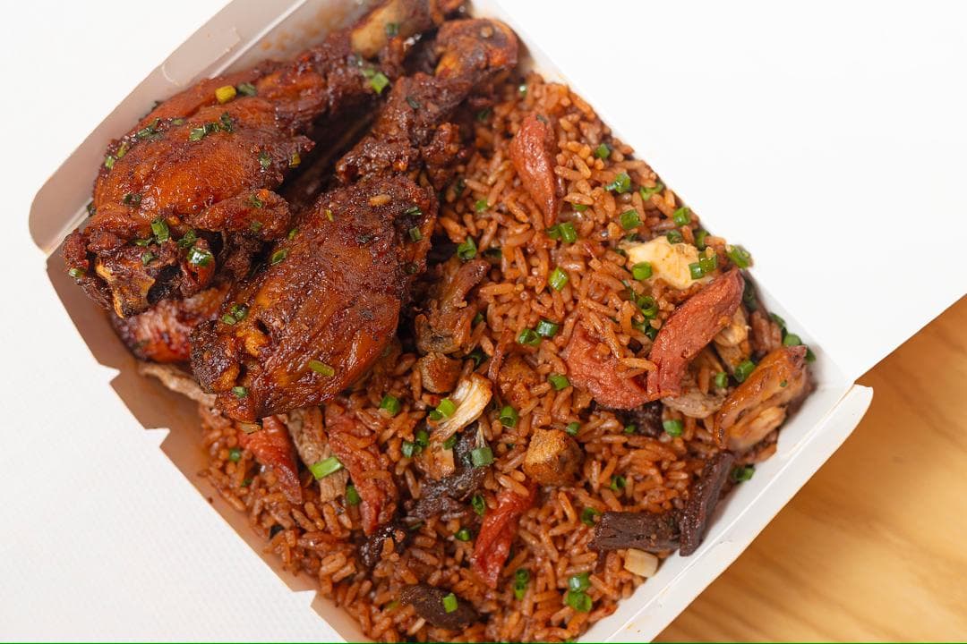 A takeaway box of jollof rice with chicken drumsticks, sausage and beef