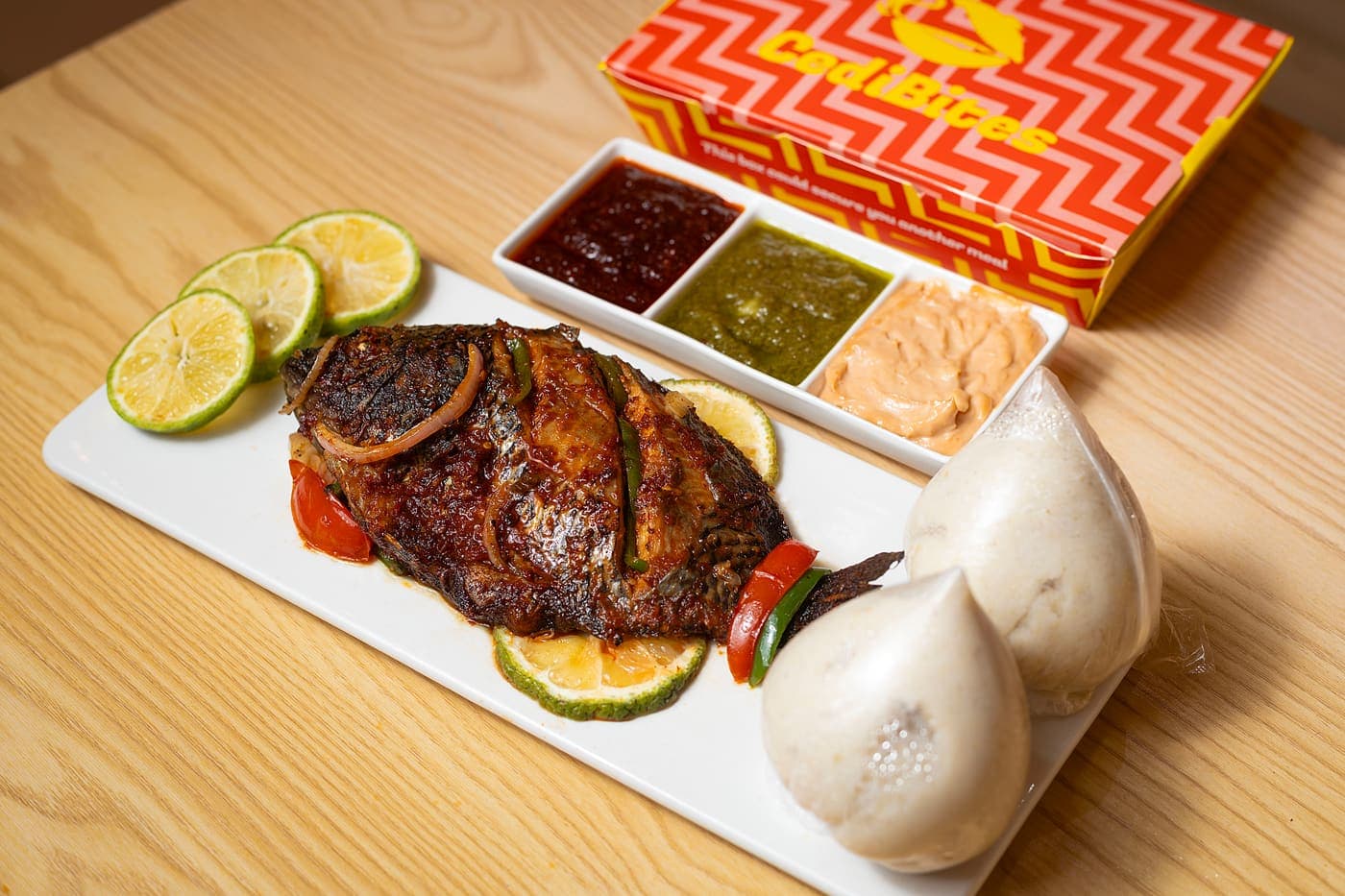 A whole grilled tilapia with lime slices on a white plate, two balls of banku beside it