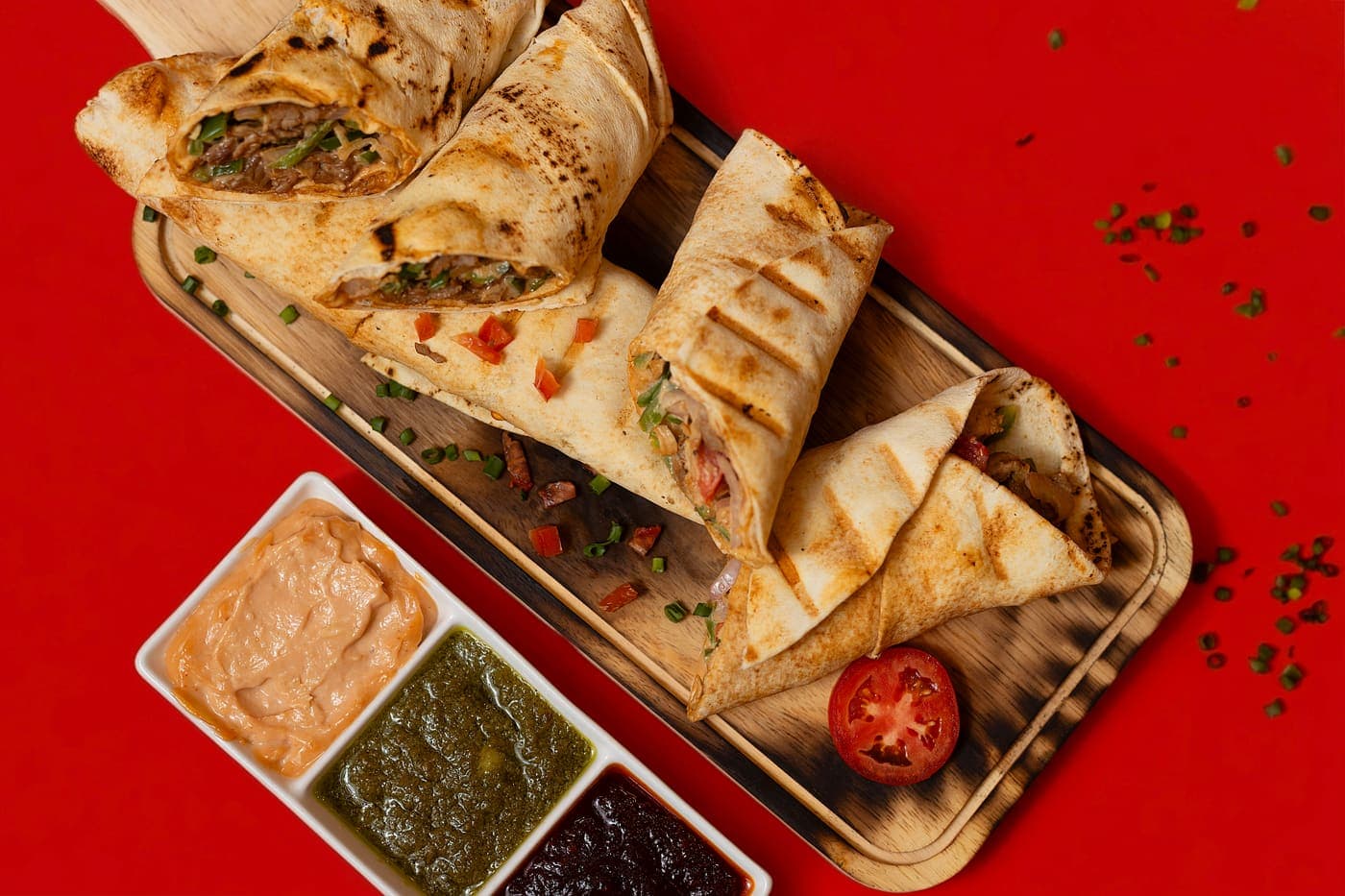 Grilled wraps cut open on a wooden board with three dipping sauces, on a red ground